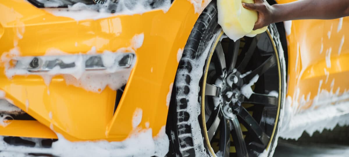 Hands of African man holding yellow sponge, washing car wheel with foam. Cleaning of modern rims of luxury yellow car at self car wash service outdoors.